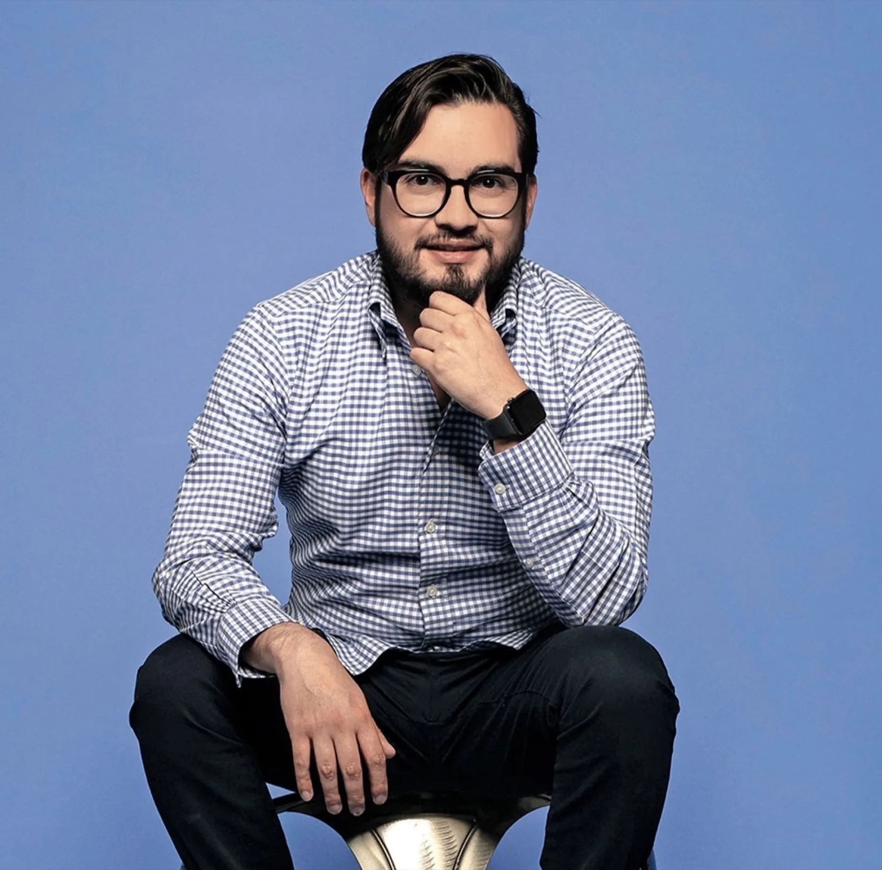 Héctor Sauza sentado, con camisa de cuadros azules y la mano en la barbilla, sobre fondo azul